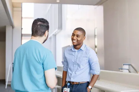 Medical professional in scrubs and office worker in a button-down shirt talk with each other.